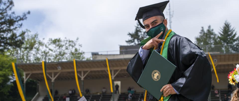 student in graduation robes and hat, holding diploma cover, in the redwood bowl