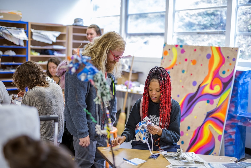 a group of students in and art classroom with a large canvas painting in the background