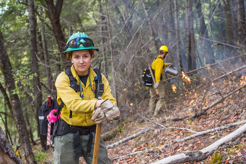 A woman in forestry gear looking into the camera leaning on an axe with a prescribed burn going on behind her